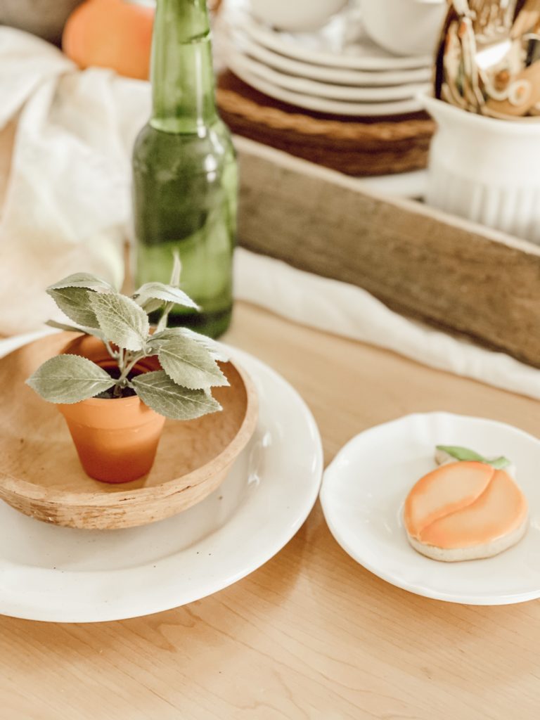 If You Love Peaches You Will Love this Tablescape plant in wooden bowl on top of white plate and a green bottle and small white plate with peach cookie on it