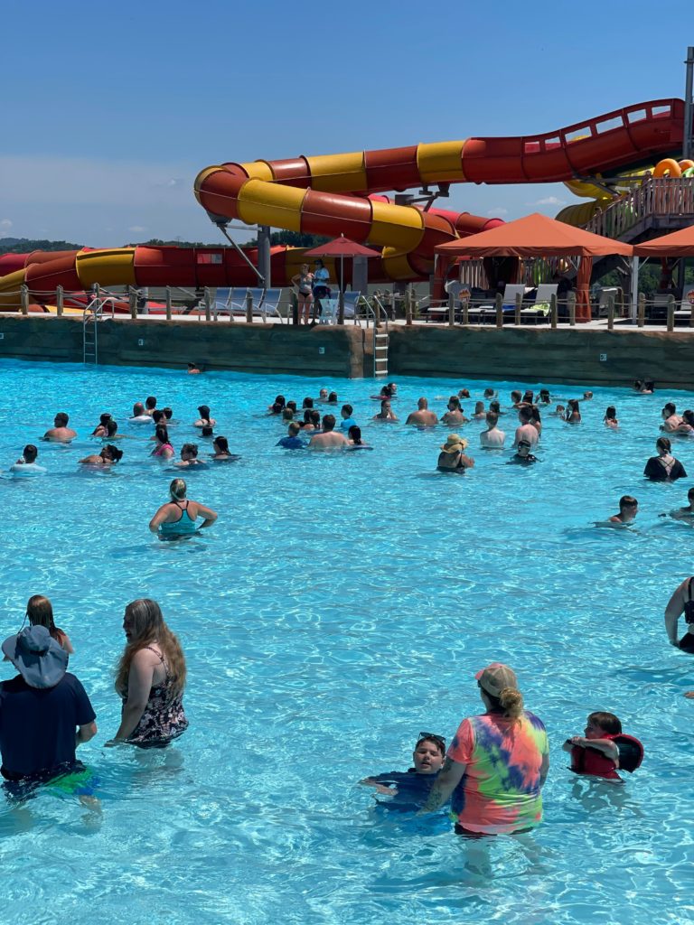 large amount of people standing in wave pool waiting for waves to start. Blue water and sky and cabanas and water slide in the background