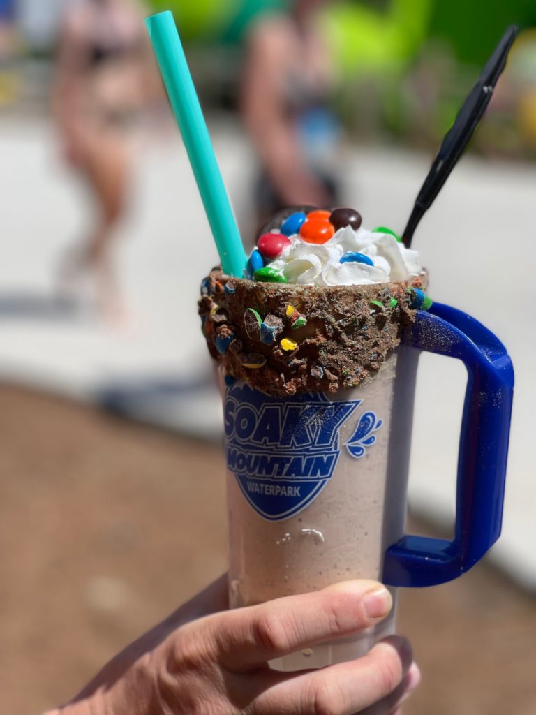 someone holding plastic cup with milk sharer chocolate candy coating around the rim with green straw and whipped cream with m&m's
