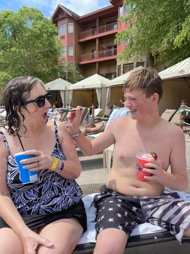 Teen boy and grandmother sitting by pool sharing a frozen drink in their swimsuits with cabanas and hotel in background