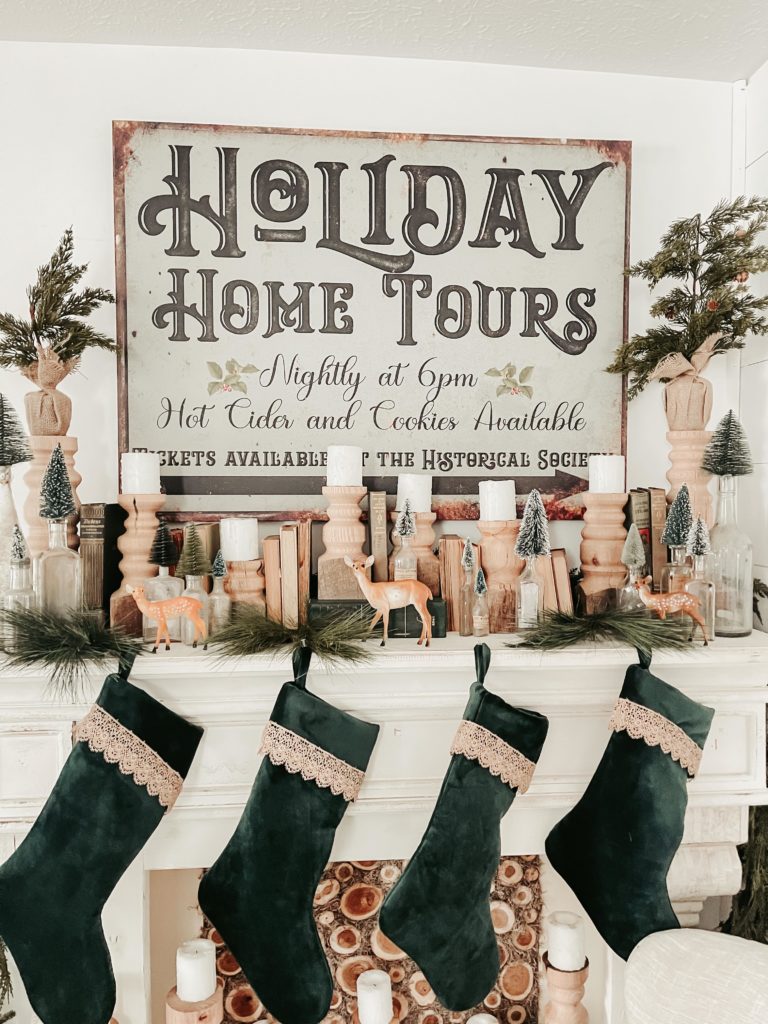 holiday home tour chippy vintage rustic looking sign on mantle with wood candlesticks and white candles green stocking and vintage bottles with bottle brush trees in them