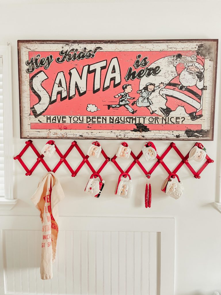 Santa is coming to town red and black sign on wall with Santa mugs hanging on rack on white wall between two windows